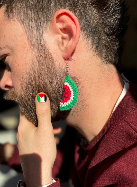 a side face profile of a male presenting individual with a full beard presents a crochet watermelon earring. he holds his face in a pensive manner, his thumb on his cheek facing the camera with a pal es tine flag painted on it